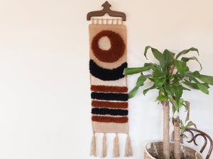 vintage tan, red and black wall weaving on a wood hook next to a house plant