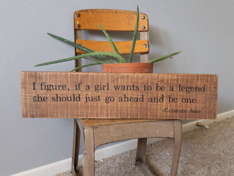 brown reclaimed wood sign with black print quoting Calamity Jane sitting on a vintage school chair along a green aloe plant